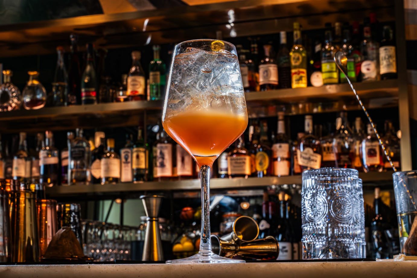 A stemmed glass on the bar, with rows of backlit bottles behind.