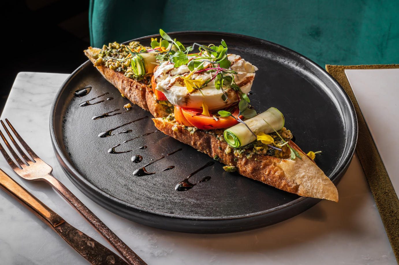 A long slice of toasted bruschetta on a dark plate, set on marble.