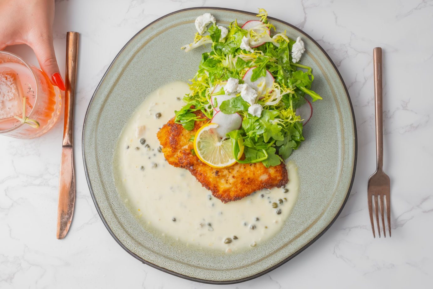 A golden chicken Milanese with dressed greens and lemon on a plate, set on marble.