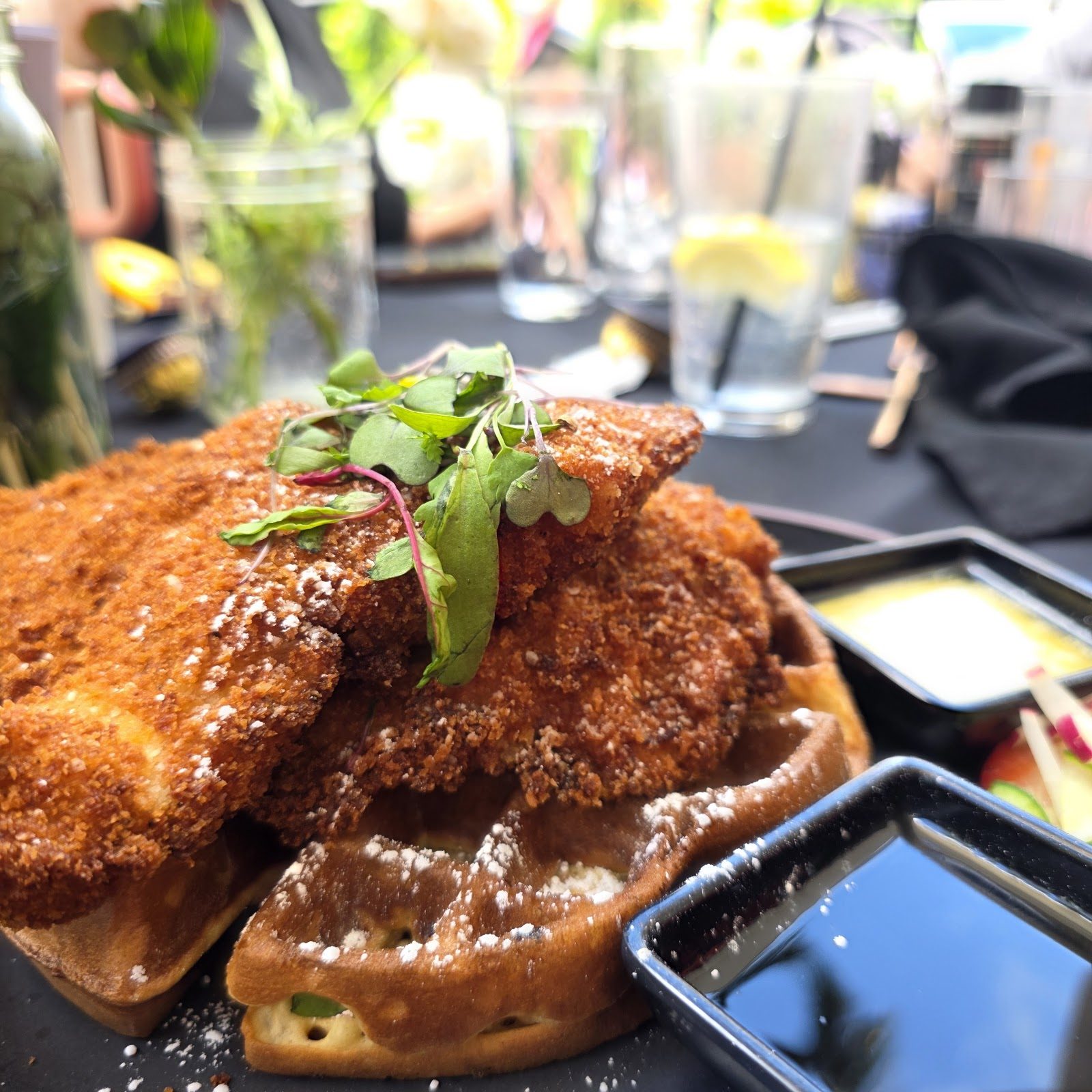 Fried chicken over a waffle, topped with microgreens, with a dipping sauce alongside.