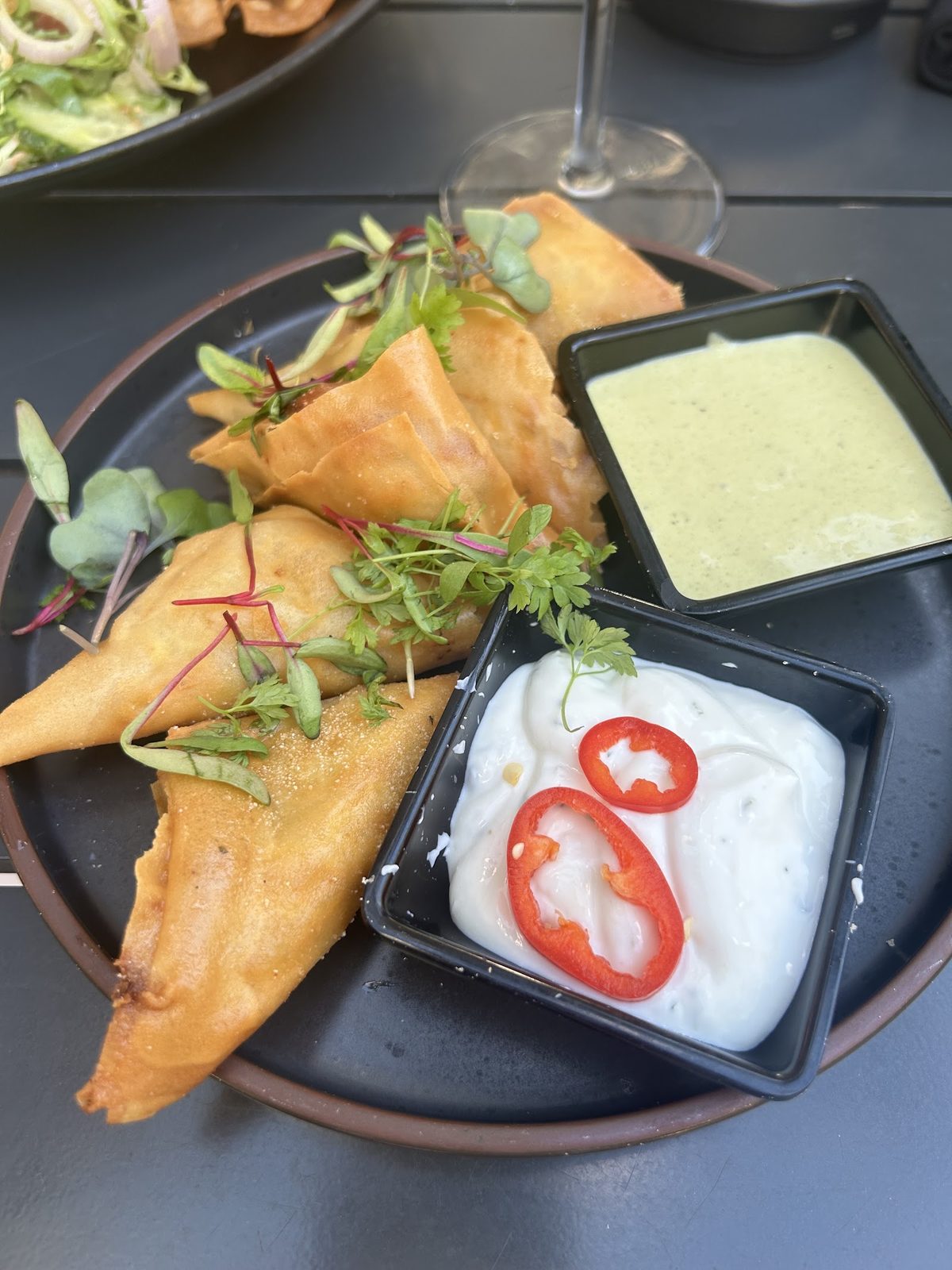 Two golden fried samosas with green and chili-flecked dipping sauces on a dark plate.