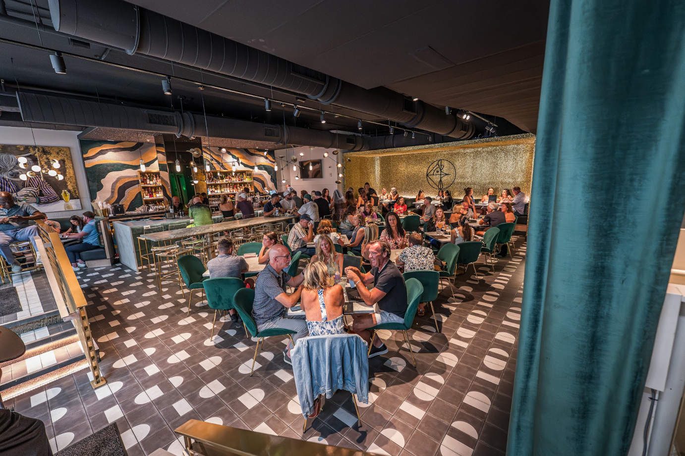 The Social Roost dining room: green velvet chairs, a checkered floor and warm light, full of guests.